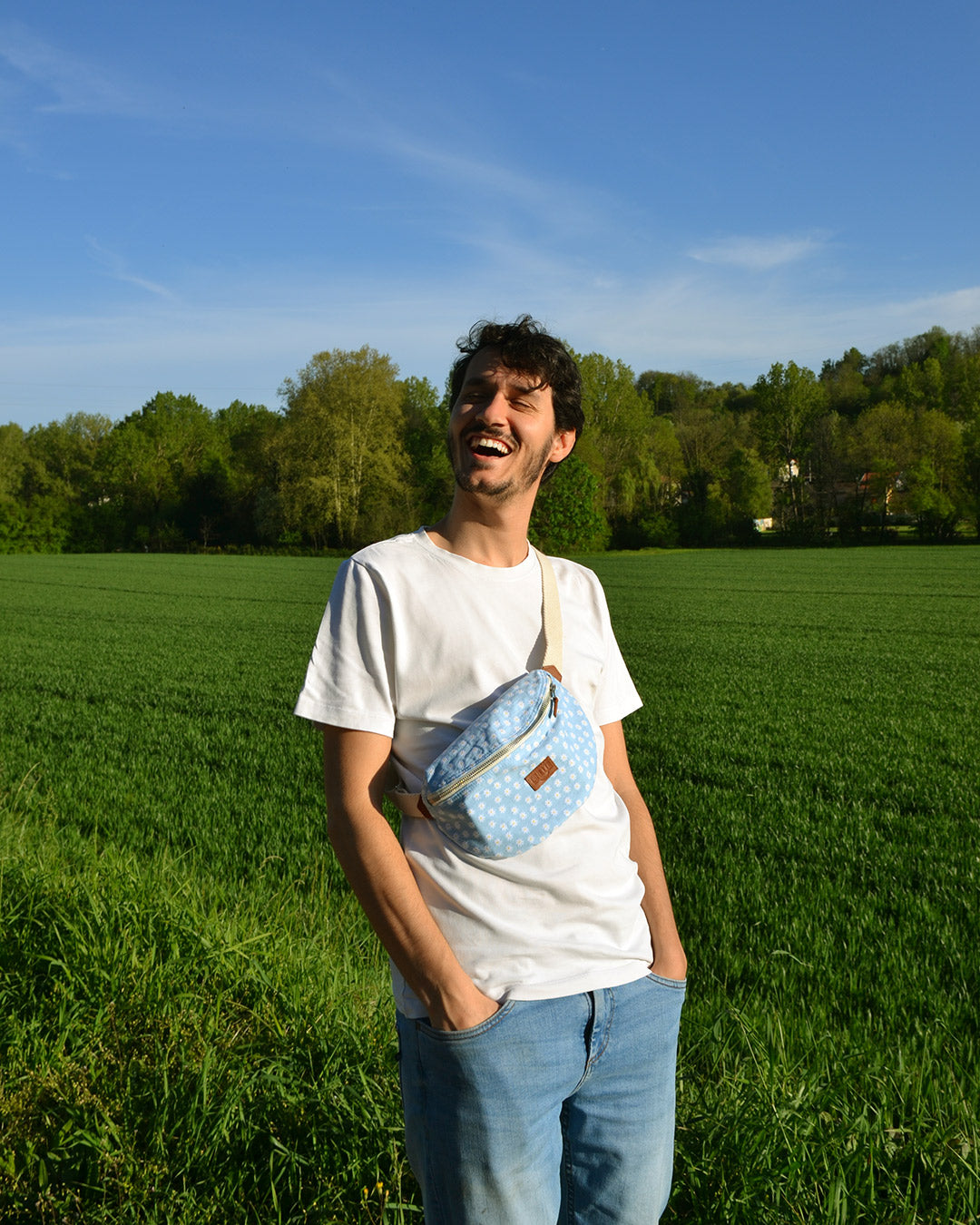 Dans cette image, un homme porte avec style un sac banane bleu orné de multiples marguerites blanches. L'accessoire ajoute une touche de fraîcheur et de couleur à sa tenue décontractée. Pratique et tendance, ce sac est parfait pour une journée en ville ou une sortie entre amis. On y retourve sur le devant le logo de Binette.